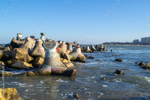 Concrete jacks painted with vibrant colors are positioned along a rocky shore, extending into the sea under a clear blue sky. The scene highlights the intertwining of art and nature.