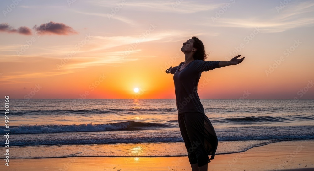 Fototapeta premium A serene woman stands on the beach at sunset with arms outstretched towards the vibrant sky