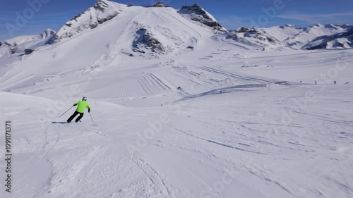 Skier in green jacket descends snowy mountain slope during sunny winter day. Slow motion POV