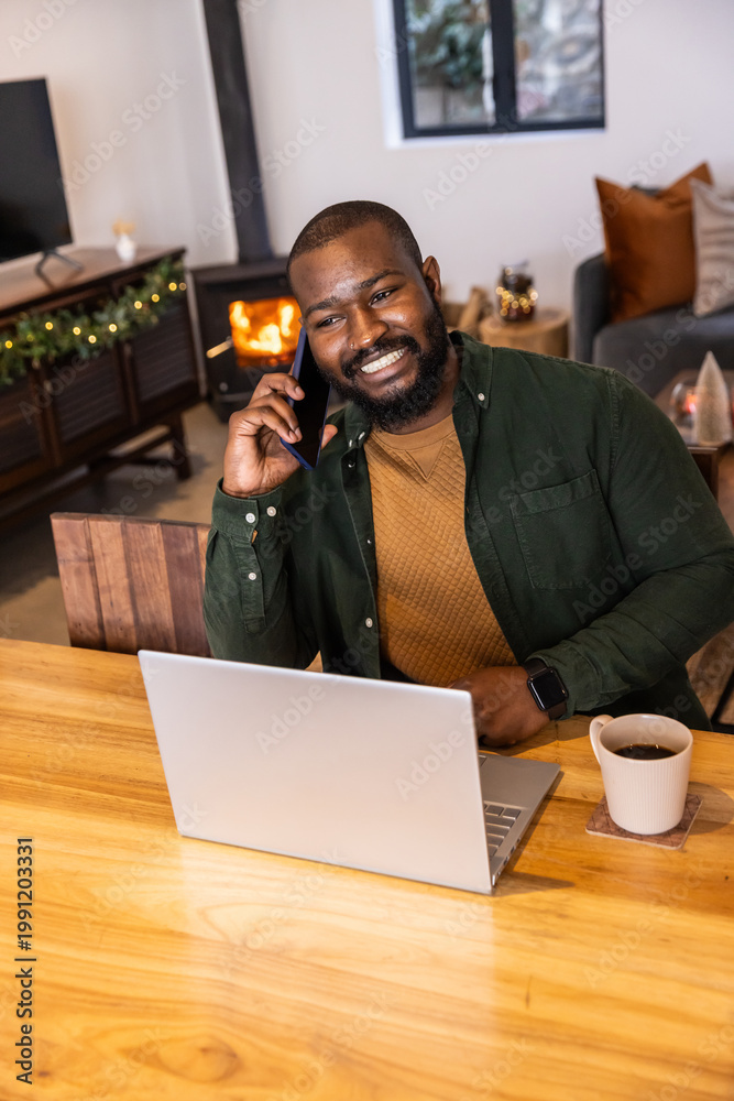 Obraz premium African American green-shirted man talking on phone while working on laptop at table in living room