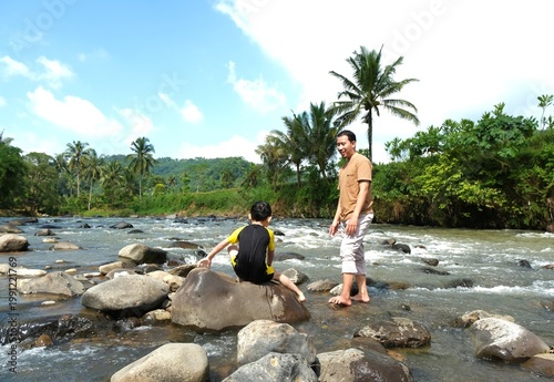 Photography Father standing and watching his son play in a shallow river in a tropical natural setting