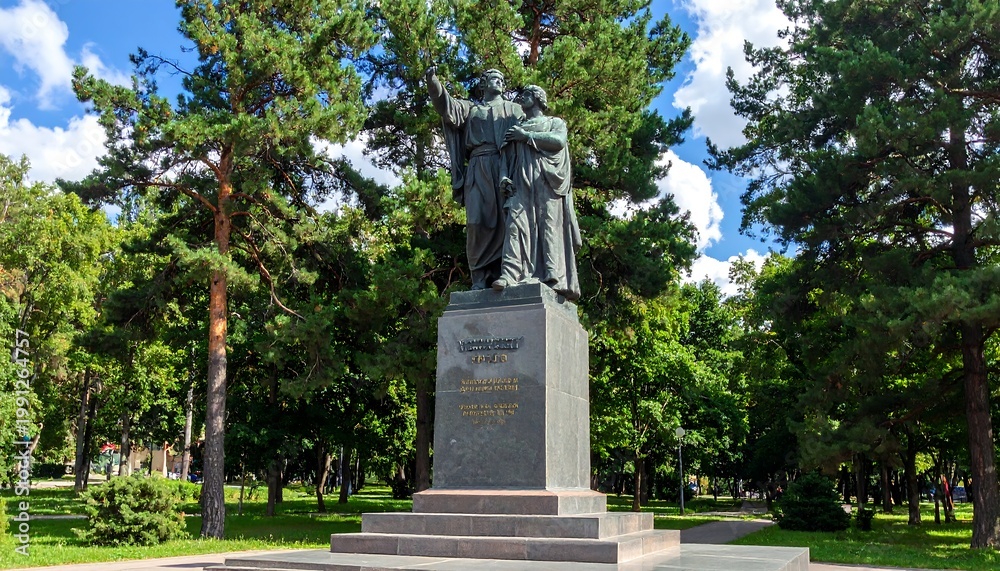 Fototapeta premium A large bronze sculpture of two figures stands atop a pedestal in a park, surrounded by lush green trees and a blue sky