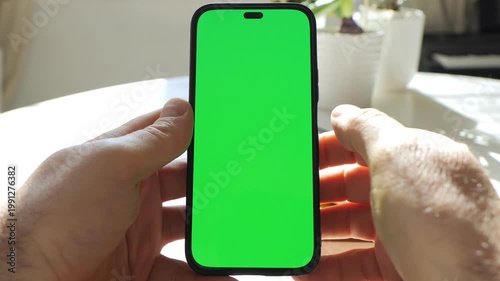 Hands hold smartphone with bright green screen. Phone centered in frame. Background shows table and white pots with plants. Sunlight creates soft shadows. Hands steady the phone without moving