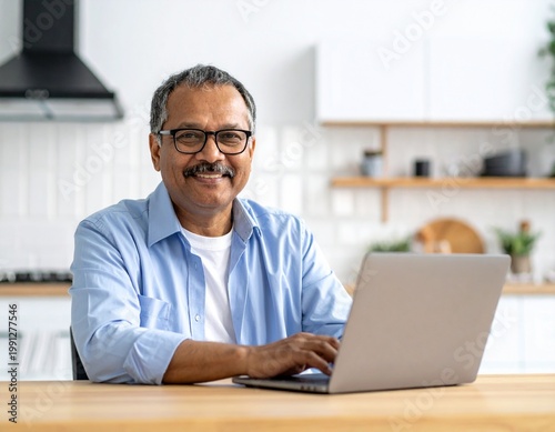 Portrait of happy middle aged senior man wearing glasses sitting with laptop at home table. Smiling older mature handsome man in eyeglasses looking at camera posing with computer sitting in kitchen.