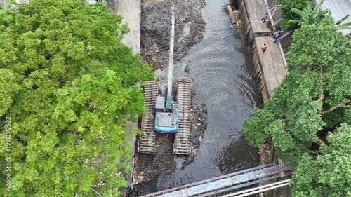 An blue excavator is working to digging the bottom of the jakarta river, Excavator with a long arm.  floating platform in a river digging cumulative dirt that shallowing the river