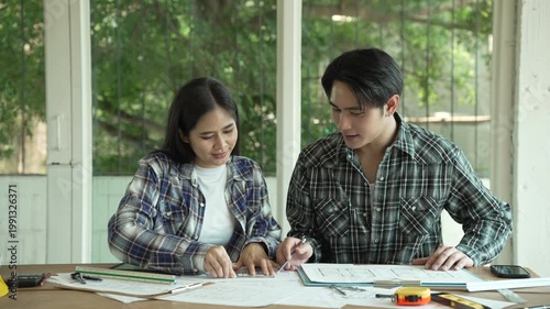 Two young adults in plaid shirts discuss paperwork at a desk in a bright room with large windows and green outdoor view, suggesting planning, collaboration, or a tense conversation.