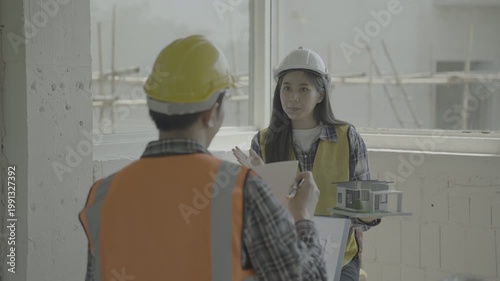Construction workers discuss building plan indoors at a site. Female engineer in hard hat holds miniature house model while colleague reviews documents. Industrial renovation, teamwork, safety.