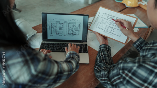 Architects reviewing house floor plans on laptop and clipboard at wooden desk, collaborative design meeting with blueprints and sketching tools in modern office.