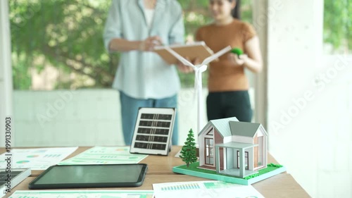 Eco-friendly home energy planning scene with model house, wind turbine and solar panel on desk, people discussing sustainability in bright office.