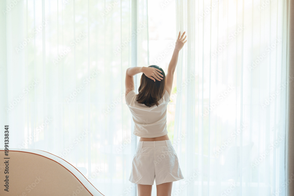Obraz premium happy woman wake up from a rest and relaxe by the window curtain, young adult female stretching, rising arms and looking to window view in the early morning. healthy lifestyle and morning routine