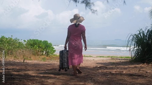 Female traveler in pink dress and straw hat approaching ocean while holding suitcase on tropical coast starting holiday journey, slow motion. Seaside travel concept