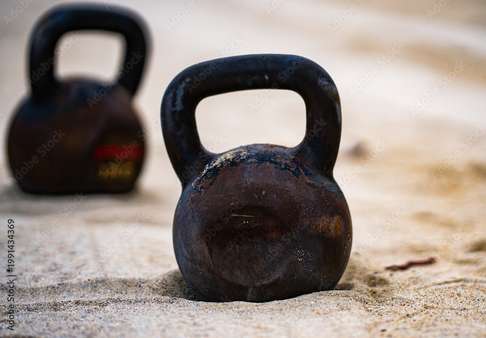 Fototapeta premium Sports equipment. Kettlebell background. Kettlebell on the beach for summer sport. Kettlebell in sand. Workout with kettlebell in public sport gym. Healthy lifestyle background. Sport banner.