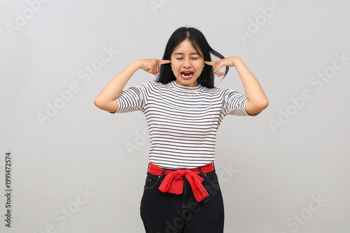 Bild auf Leinwand A young woman stands isolated on a white background, looking angry, covering her ears with her fingers and screaming