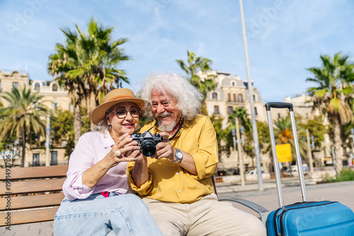 Joyful senior couple examining photos on a vintage camera while relaxing on a bench during their city vacation