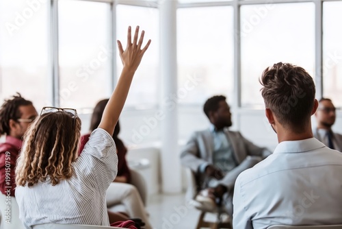 A woman raises her hand to ask a question during a diverse group discussion in a bright, modern office setting.