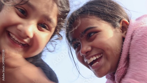 Two young Indian girls smile brightly while looking down toward the camera, capturing a joyful and playful childhood moment with natural expressions in a simple rural village setting