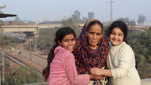 An elderly Indian grandmother stands with her two granddaughters, all smiling warmly together, capturing a joyful moment of family bonding and love in a peaceful rural village environment