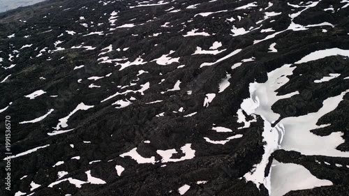 Aerial view of volcanic landscape with black lava rock and patches of white snow, showcasing the rugged terrain and natural formations in a remote area