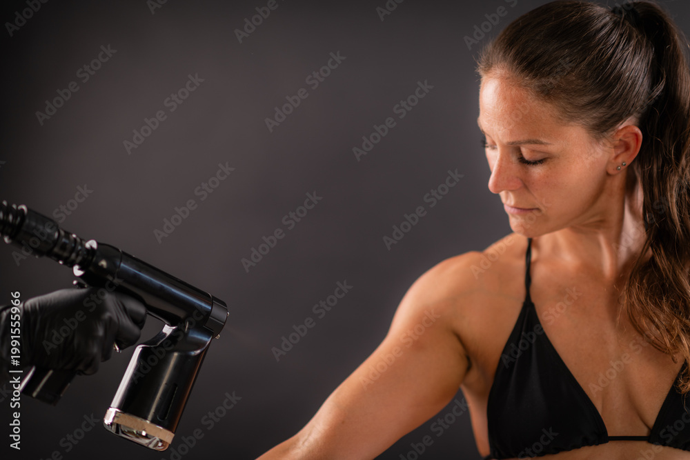 Fototapeta premium Applying a fine tanning mist to the upper arm using a professional spray device. Controlled technique supports even coverage and uniform cosmetic skin tone.