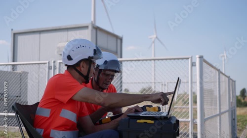 Asian engineers using a laptop to monitor wind turbine data at a renewable energy site, representing clean energy operations and digital analysis in sustainable engineering.
