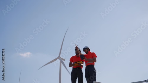 Asian engineers using a tablet and clipboard to inspect wind turbines at a renewable energy site, representing clean energy operations and technical teamwork in sustainable engineering.