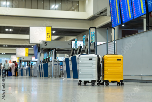 Two modern hard-shell suitcases, yellow and white, are standing in a blurred airport terminal departure hall near check-in counters and flight information displays, ready for travel and vacation.
