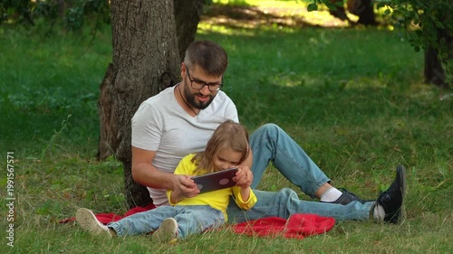 Happy family father and little girl sit on green lawn in summer park with digital tablet. Computer games surfing internet social media. Picnic outdoors