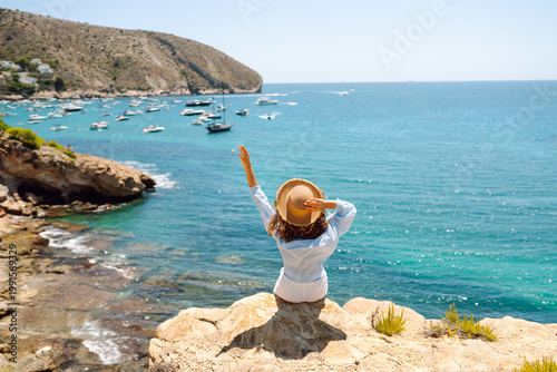 Fotografía Rear view of a young woman in a hat sitting on the edge of a cliff overlooking the sea