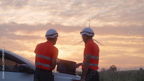 Asian engineers analyzing wind turbine data on a laptop at sunset, representing renewable energy monitoring, digital analysis, and teamwork in sustainable energy operations.