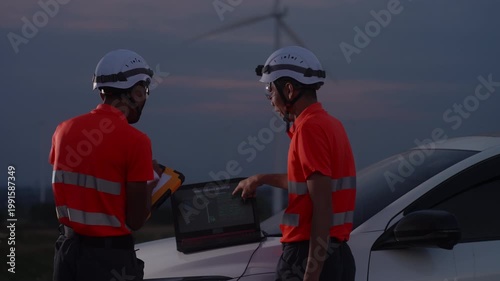 Asian engineers analyzing wind turbine data on a laptop at an evening renewable energy site, representing digital monitoring, clean energy operations, and teamwork in sustainable engineering.