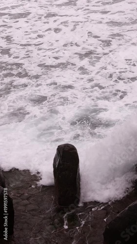 Vertical high-angle shot of white ocean foam and powerful waves swirling around dark volcanic rocks on a moody, overcast day