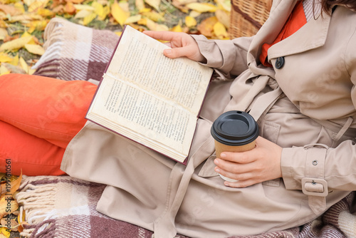 Young woman with paper cup of coffee reading book on plaid in autumn park