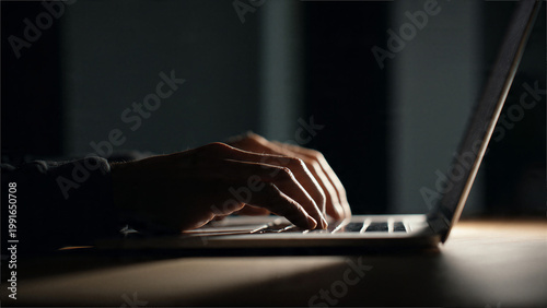 Dark workspace with hands typing on laptop illuminated by screen light