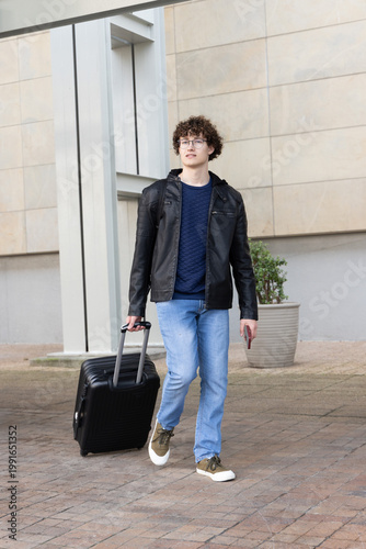 Adult man walking on plaza by column, pulling black suitcase, holding phone, wearing glasses
