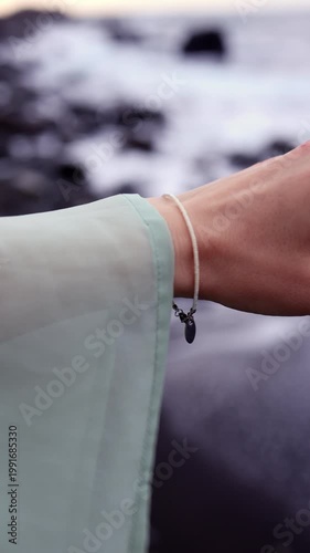 A vertical close-up shot of a woman's hand wearing a delicate silver bracelet with a pearl, with a blurred rocky beach and ocean waves in the background