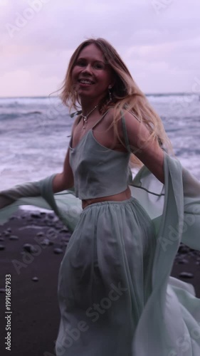A joyful vertical shot of a blonde woman laughing and spinning on a dark volcanic beach, with the wind blowing her flowing mint-green outfit against a backdrop of ocean waves