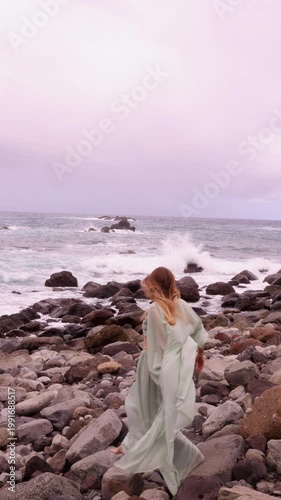 Vertical back view of a woman with long wavy hair, wearing a flowing mint-green outfit, walking over dark coastal rocks towards powerful ocean waves under a moody, overcast sky