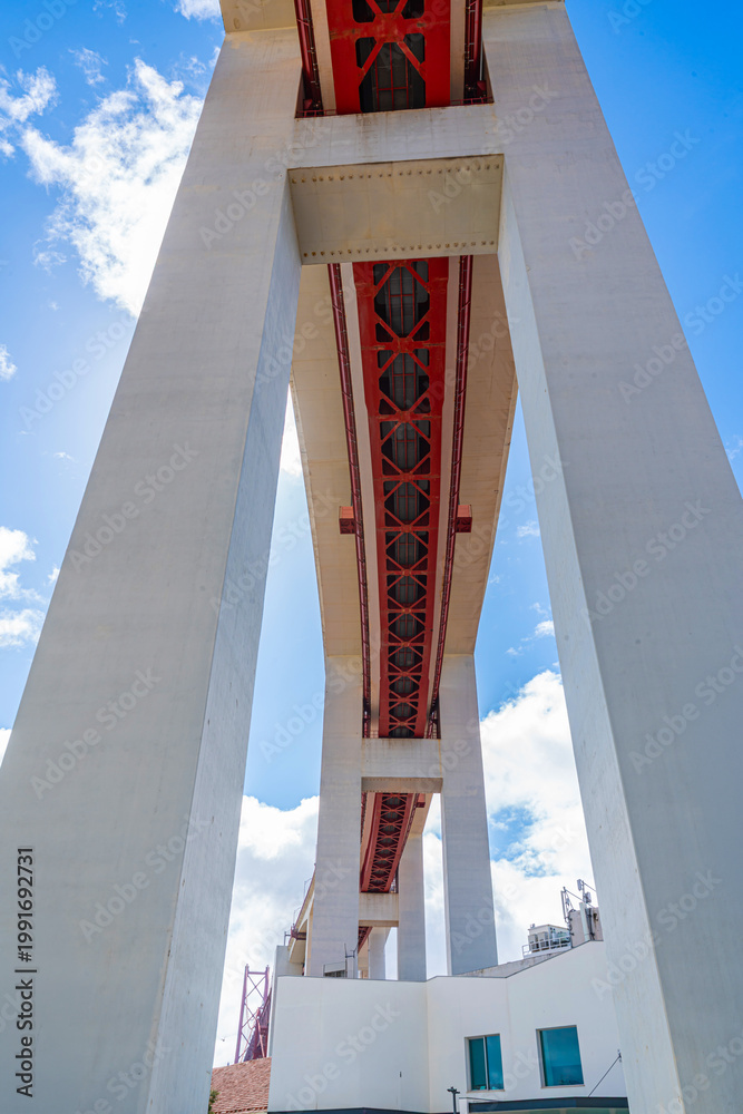 Fototapeta premium Low angle view from beneath a large modern bridge showing massive concrete pillars and red steel framework against blue sky with clouds.