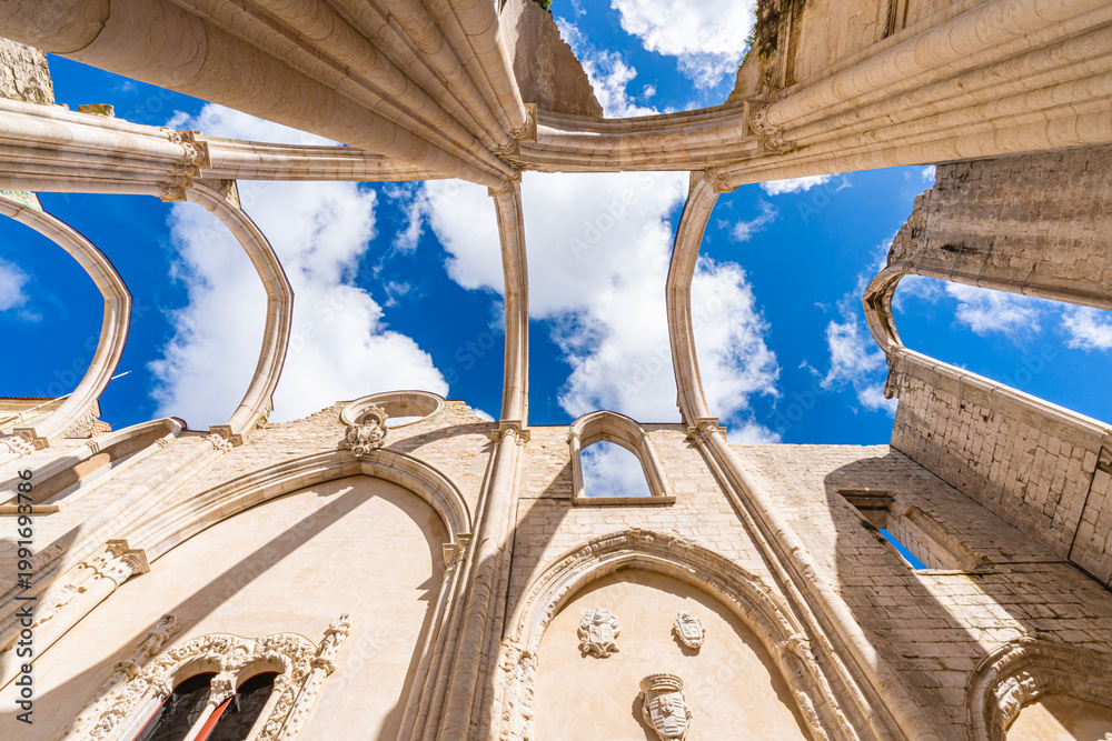 Fototapeta premium Dramatic upward perspective of Carmo Convent ruins in Lisbon with gothic arches and columns framing blue sky, highlighting historic architecture and open roof structure.