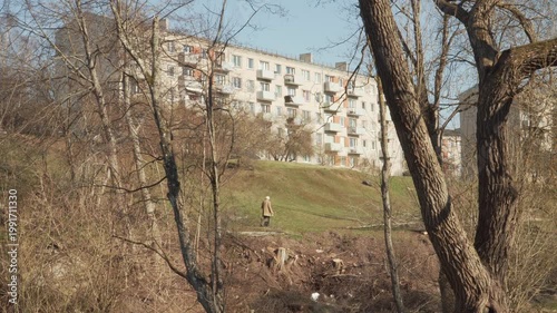 A senior woman walks her dog across a grassy slope on a sunny spring day. Bare trees frame a multi-story apartment building in a quiet residential area during the early afternoon. 