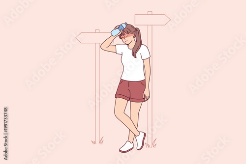 Female trekker standing near direction signs in summer heat. Exhausted girl cooling off with water bottle during mountain hike at road crossing.