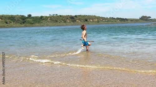 Carefree young boy wearing blue swim trunks running and joyfully playing with a wooden stick in the shallow water of a river on a beautiful sunny day, splashing around on the sandbar