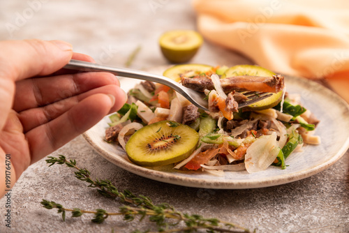 Meat salad with beef, kiwi and vegetables with hand on brown concrete, side view, close up, selective focus