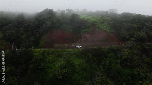 Drone Clip of white Car driving up a mountain during monsoon 4K60. The shot is moving away from the camera as the car moves ahead. 4K60fps