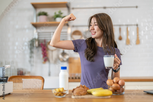Fitness happy asian woman flexing bicep holding glass of milk in kitchen. Smiling female having healthy breakfast, protein diet and calcium for workout nutrition. Healthy active lifestyle concept