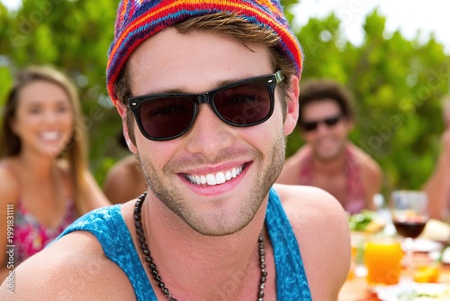 A smiling man wearing sunglasses and a colorful hat with friends in the background