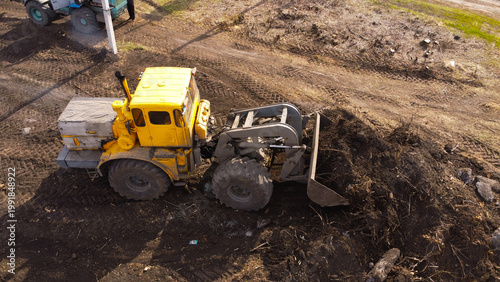 A high-angle aerial photograph of a classic yellow heavy-duty tractor equipped with a front loader bucket is Clearing Dirt at Construction Site