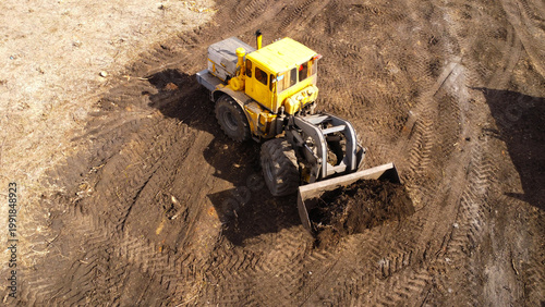 A high-angle aerial photograph of a classic yellow heavy-duty tractor equipped with a front loader bucket is Clearing Dirt at Construction Site