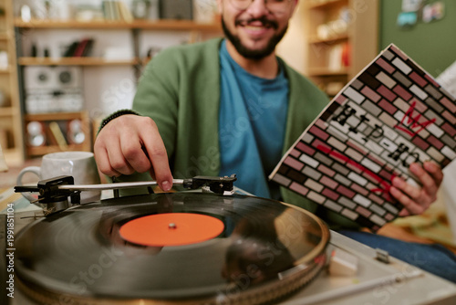 Foto Young adult man playing vinyl record on turntable while holding album cover, enj
