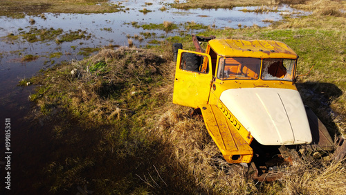 aerial shot of an old, rusted orange and white heavy-duty truck cabin and chassis abandoned in a swampy, waterlogged field